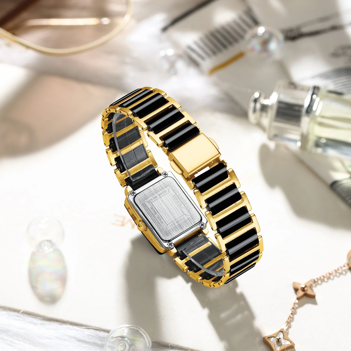 Gold and black rectangular watch on a reflective surface with blurred jewelry and perfume bottles in the background