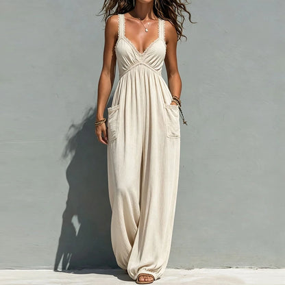 A woman wearing a cream-colored sleeveless maxi jumpsuit with lace trim, side pockets, and a gathered waist, standing against a textured gray wall.