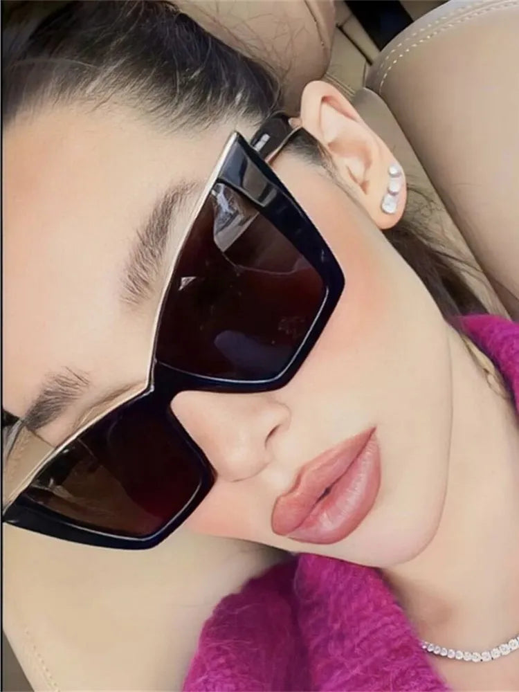 A close-up selfie of a person wearing oversized square sunglasses with a glossy black frame and dark lenses, resting against a beige car seat.