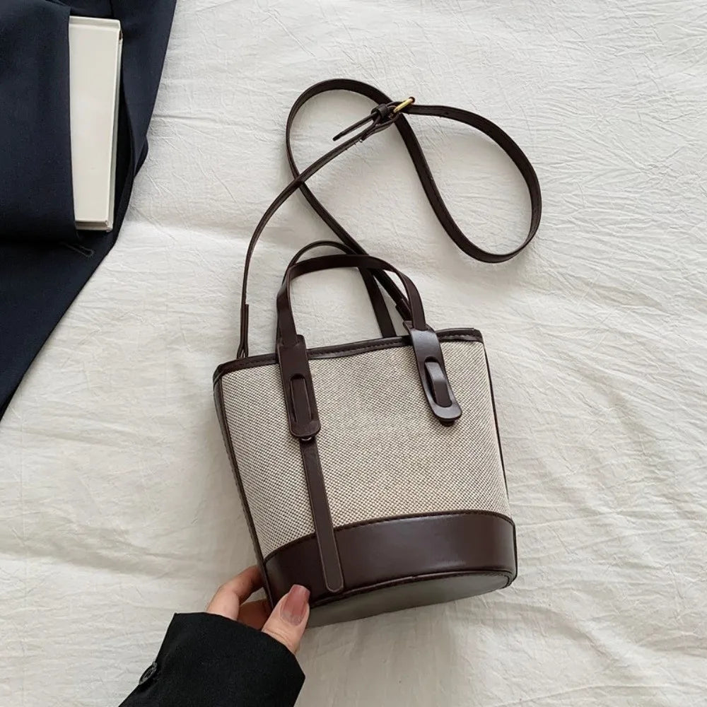 A beige woven bucket bag with dark brown leather trim, top handles, and a long shoulder strap, held by a hand over a white textured fabric background.