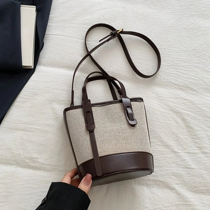 A beige woven bucket bag with dark brown leather trim, top handles, and a long shoulder strap, held by a hand over a white textured fabric background.
