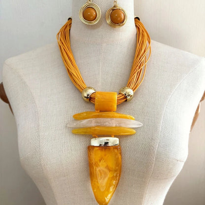 A statement necklace and matching drop earrings displayed on a cream fabric mannequin torso. The necklace features multiple strands of orange cord, gold-toned metal beads, stacked translucent amber and white stone pieces, and a large polished amber pendant.