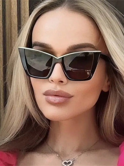 A close-up of a woman wearing black cat-eye sunglasses with a thin gold metal frame and dark lenses, paired with a silver heart pendant necklace.