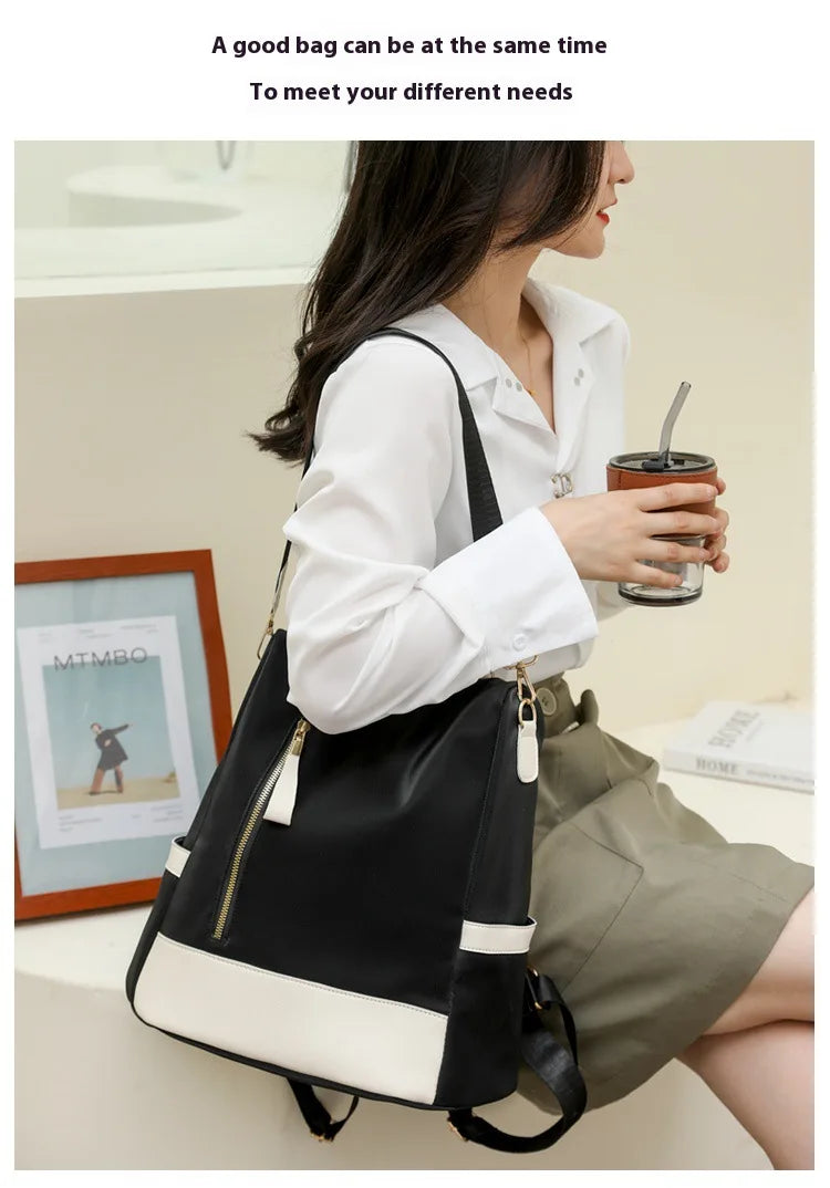 A woman wearing a white shirt and khaki skirt carries a black and white shoulder bag with gold zipper details while holding a drink. The photo is set in a bright indoor environment with a framed picture on the wall.