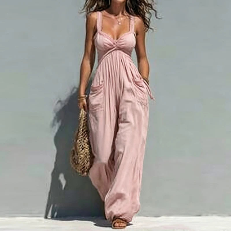 A woman wearing a light pink sleeveless maxi dress with a gathered waist and wide-leg pants, standing against a pale blue-gray wall and holding a woven straw bag.