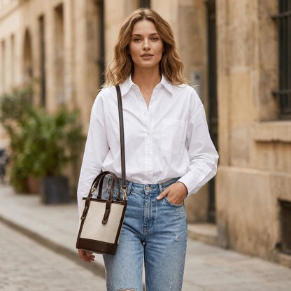 A woman wearing a white button-down shirt and light blue jeans walks down a cobblestone street, carrying a beige and brown crossbody handbag.