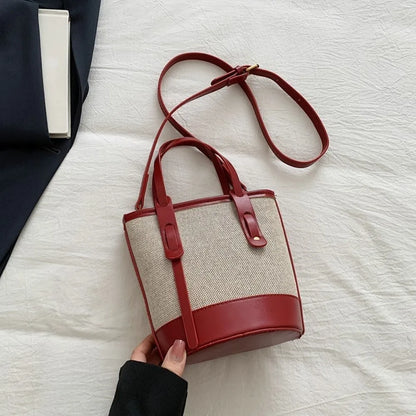 A red and beige woven bucket bag with a long shoulder strap, held by a hand over a white textured fabric background.