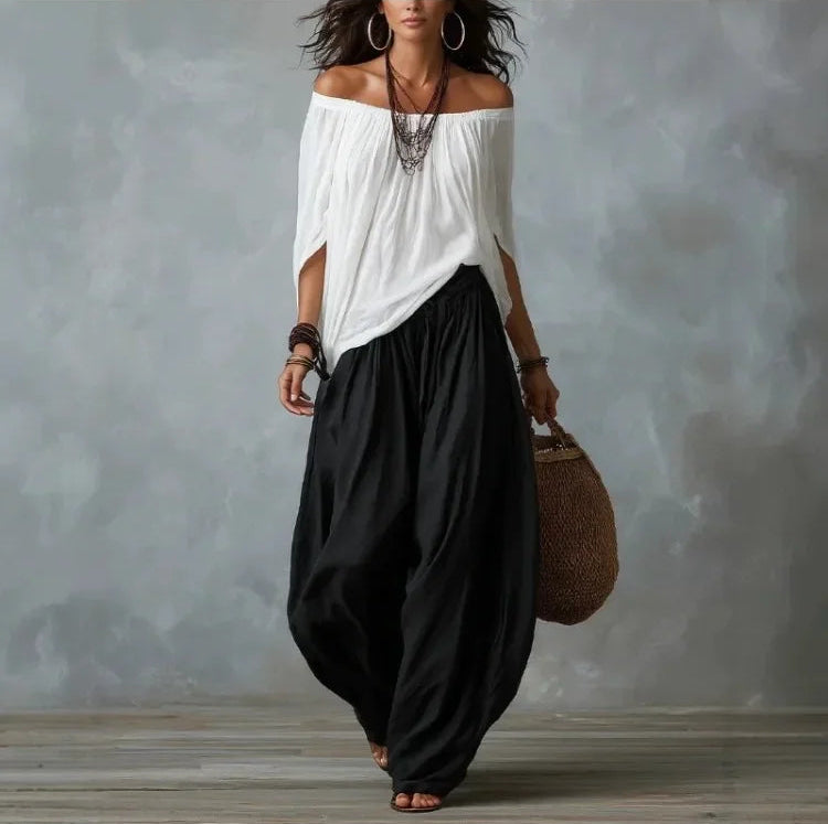 Woman wearing a white off-shoulder top and black harem pants against a gray background