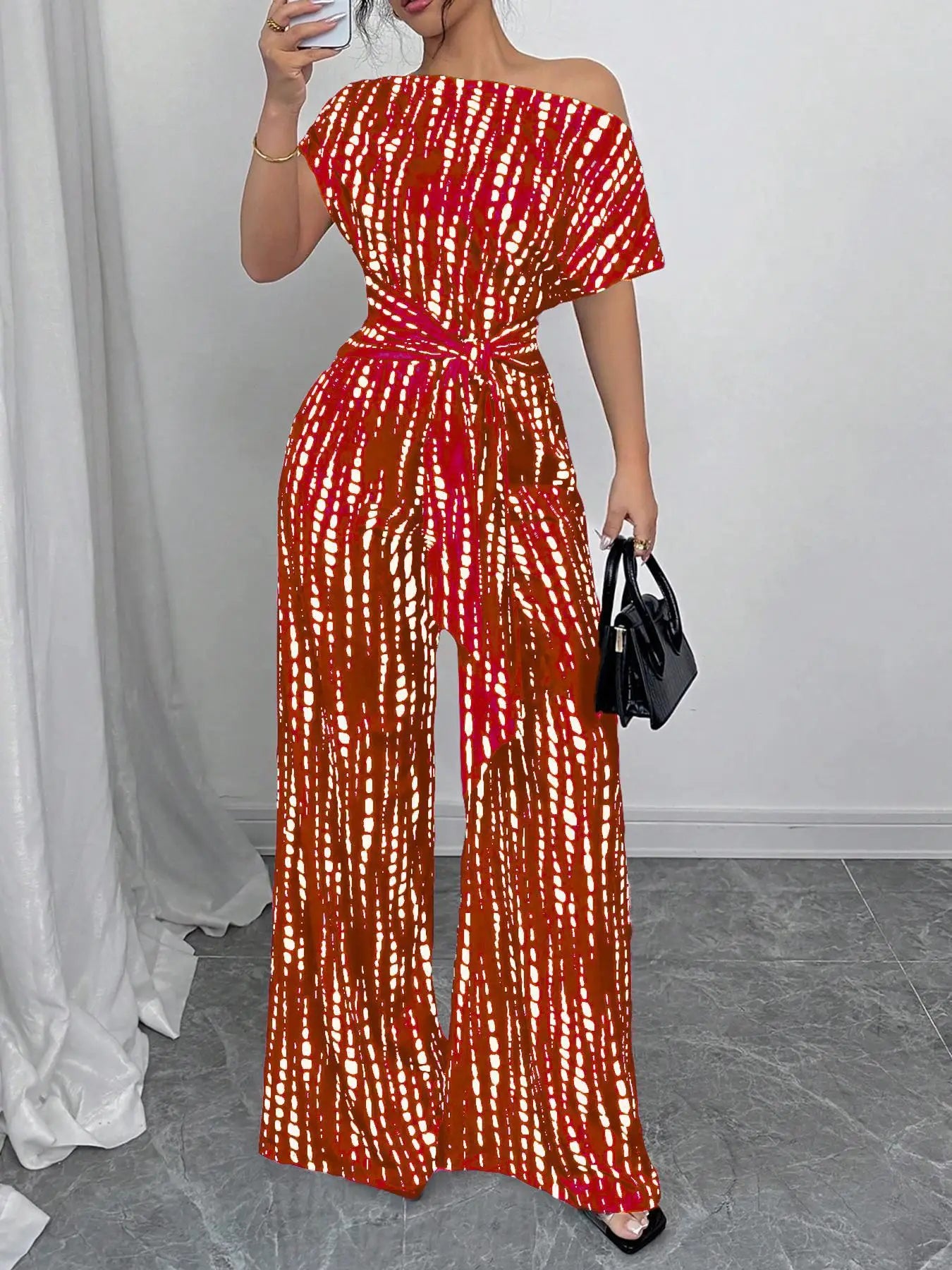 A woman modeling a one-shoulder off-the-shoulder jumpsuit with a red, orange, and white tie-dye pattern. She is holding a black handbag and standing in front of a white curtain and gray tiled floor.