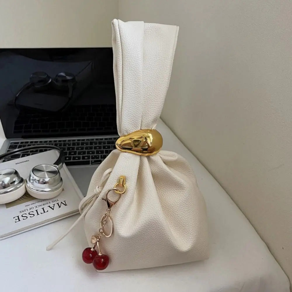 White drawstring bag with gold accents and red charms on a surface with a laptop and magazine in the background.