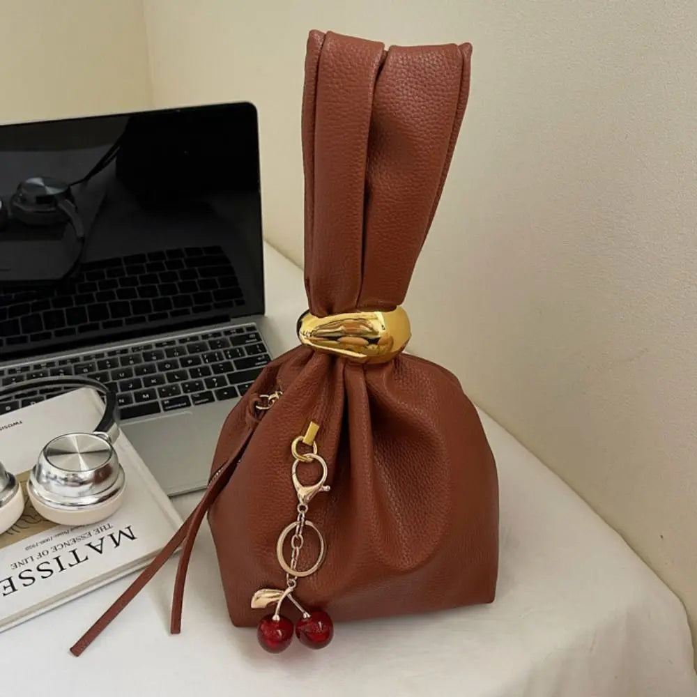 Brown leather drawstring bag with gold accents and cherry charms on a white surface next to a laptop.