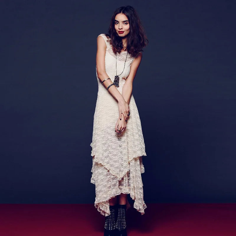 A woman modeling a sleeveless cream lace midi dress with an asymmetrical ruffled hem, styled with layered necklaces and studded black ankle boots against a dark studio backdrop.