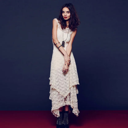 A woman modeling a sleeveless cream lace midi dress with an asymmetrical ruffled hem, styled with layered necklaces and studded black ankle boots against a dark studio backdrop.