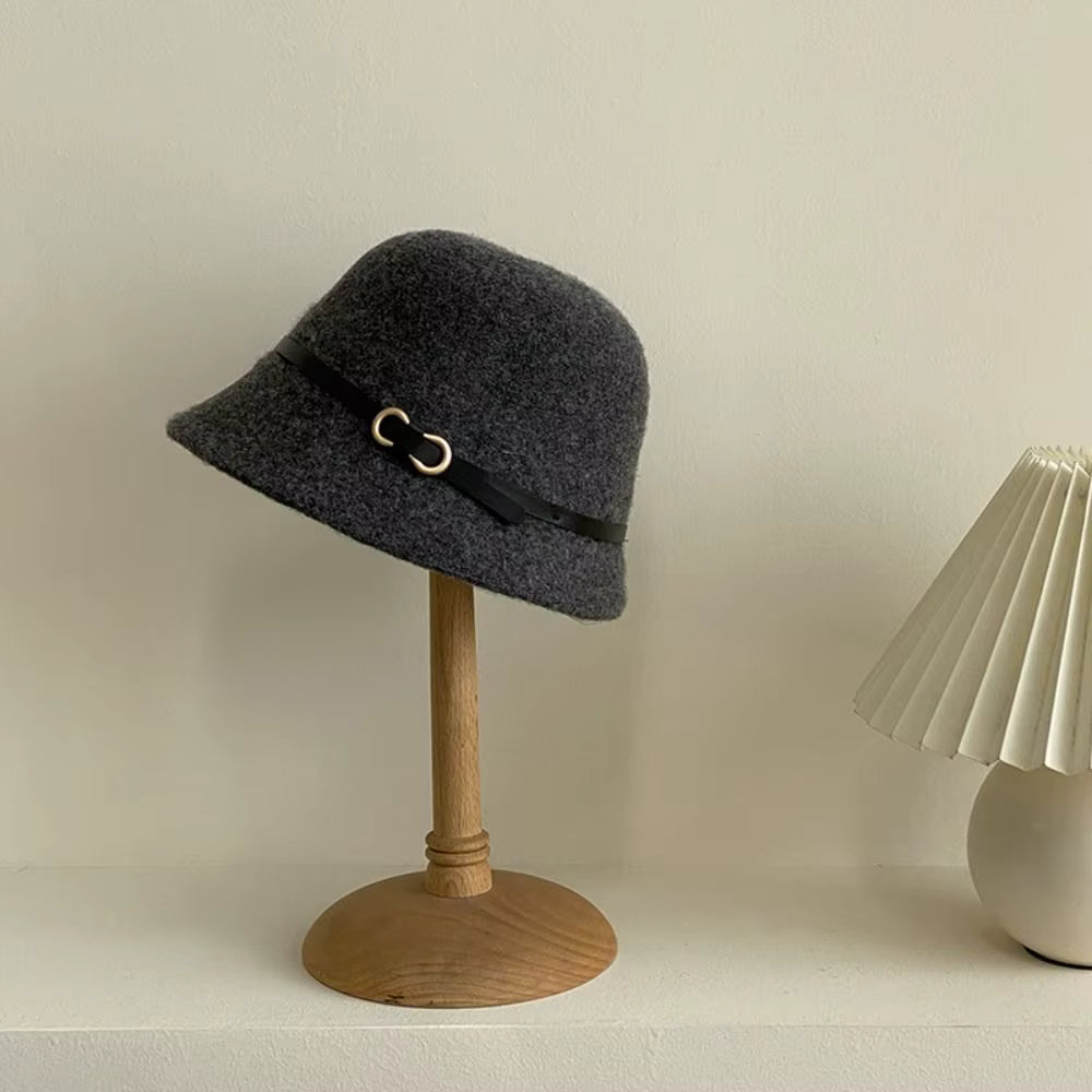 A dark gray felt bucket hat with a black ribbon band and gold-tone buckle, displayed on a wooden stand against a warm off-white wall.
