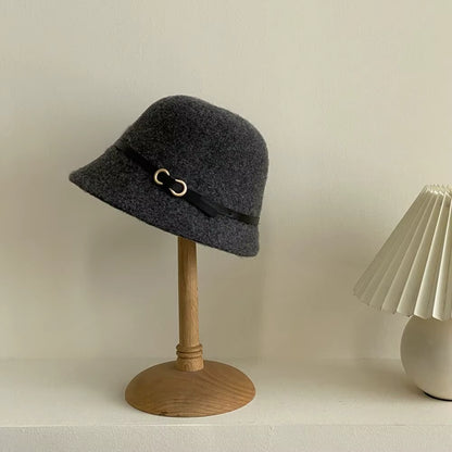A dark gray felt bucket hat with a black ribbon band and gold-tone buckle, displayed on a wooden stand against a warm off-white wall.