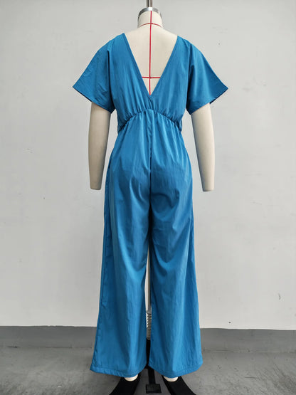A bright blue short-sleeve jumpsuit shown from the back on a mannequin, featuring a deep V-neckline and wide legs, set against a plain light gray wall.
