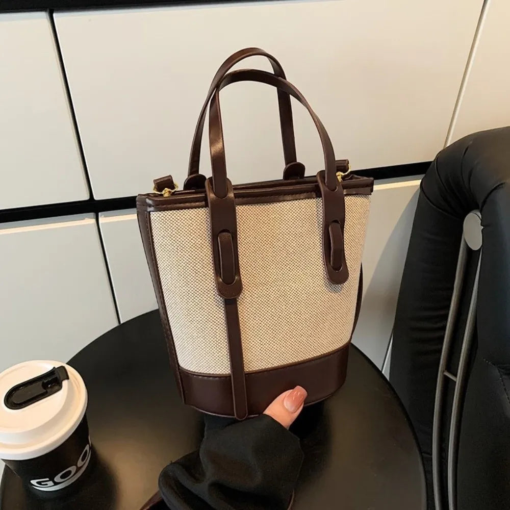A beige woven bucket bag with dark brown leather trim and handles, held by a hand over a black table next to a coffee cup.