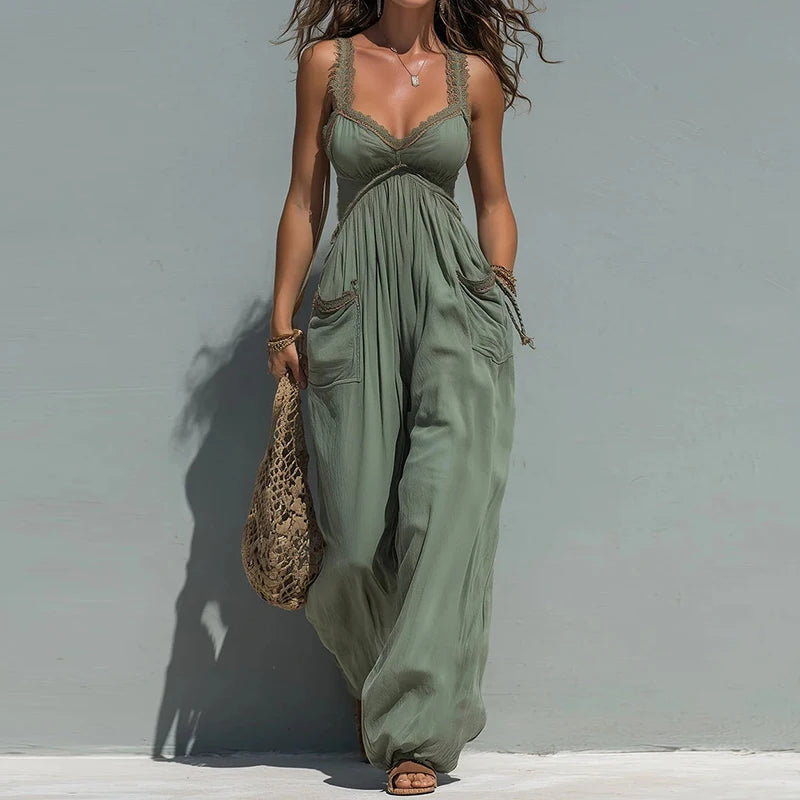 A woman models a sage green sleeveless jumpsuit with lace trim and side pockets, holding a woven straw bag against a plain gray wall.
