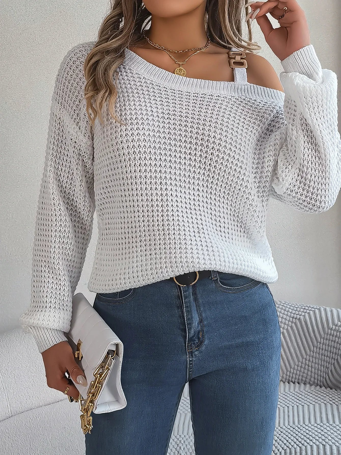 Woman wearing a white off-shoulder sweater and blue jeans, holding a white clutch with gold accents.