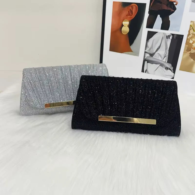 Two glittery clutch bags, one black and one silver, with gold-tone bar clasp closures, displayed on a white faux fur surface with a fashion magazine in the background.