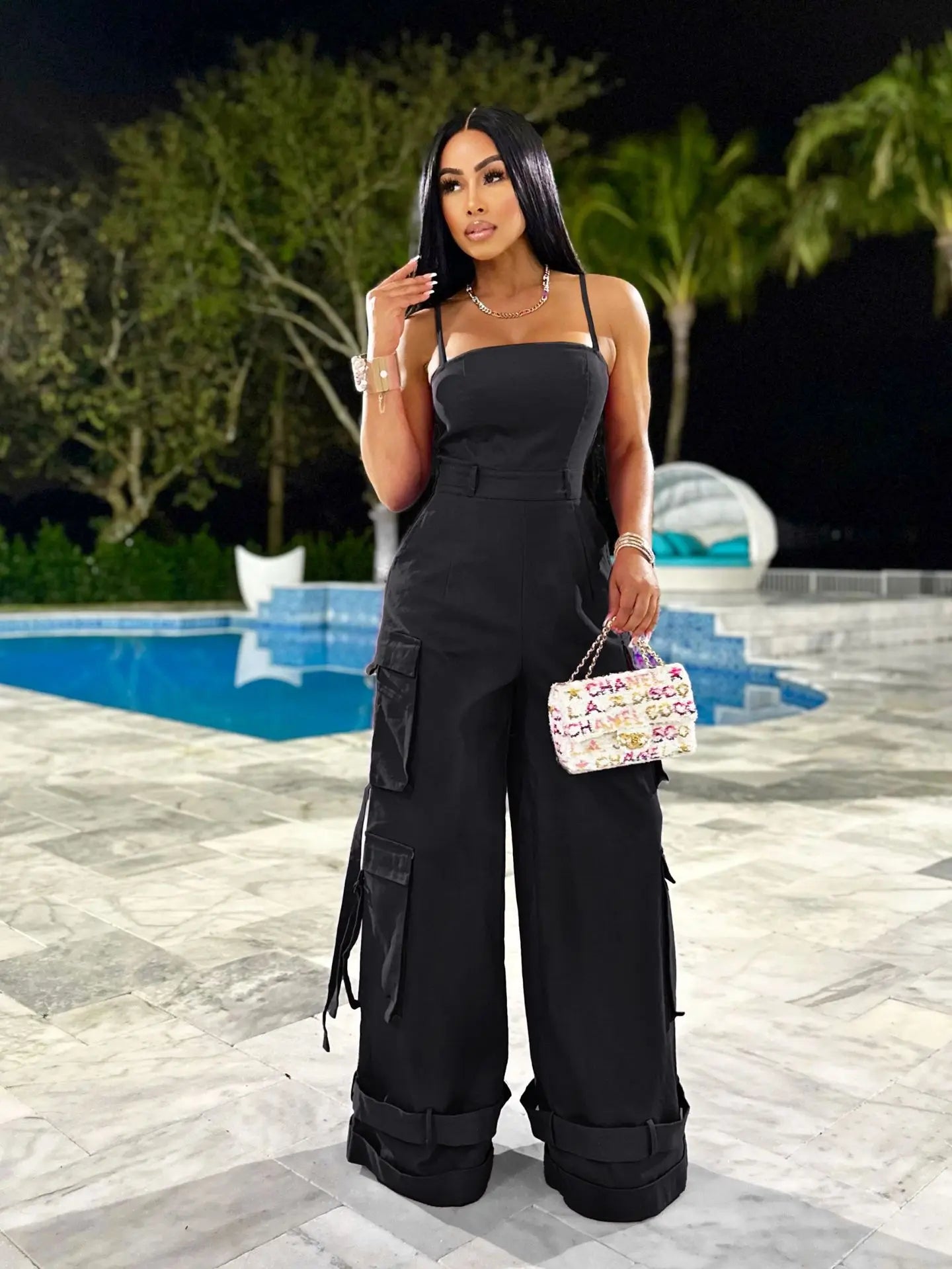 Woman in black jumpsuit holding a colorful handbag by a pool at night.