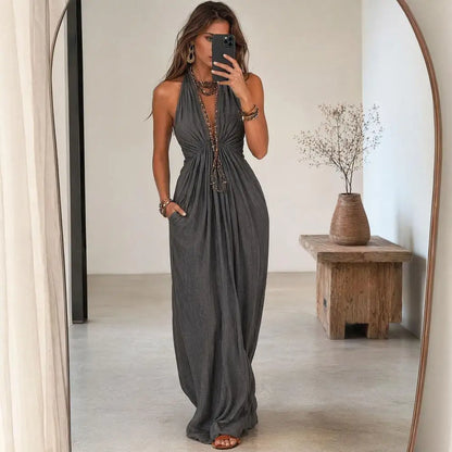 A woman takes a mirror selfie wearing a dark gray halter-neck maxi dress with a deep V-neckline, side pockets, and a gathered waist. The setting is a minimalist room with a wooden side table and a large arched mirror.