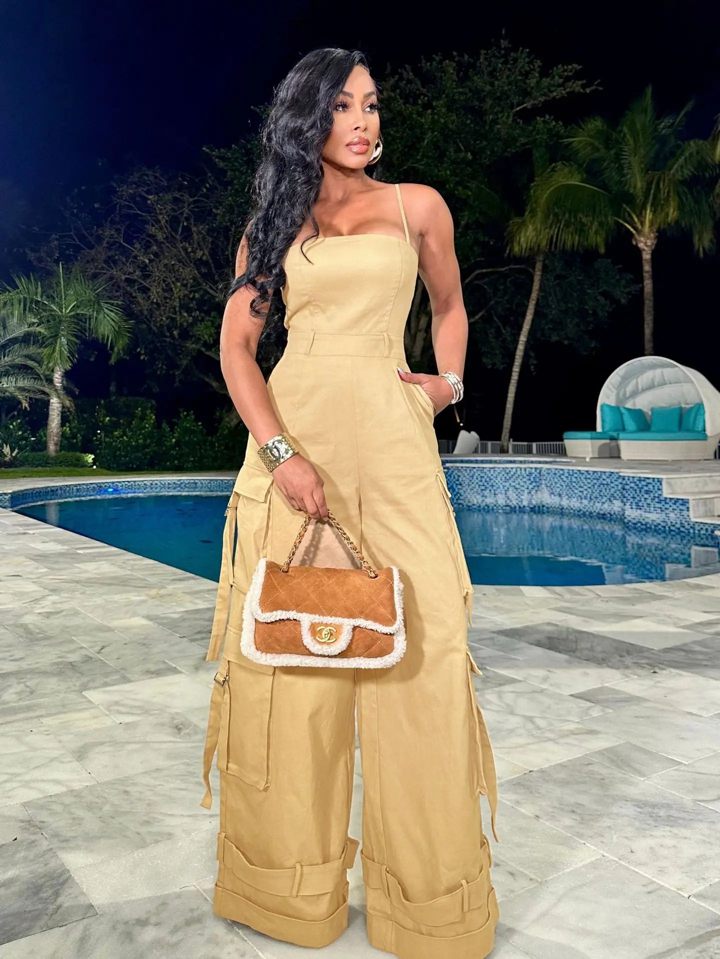 Woman in a beige outfit holding a handbag by a pool at night.