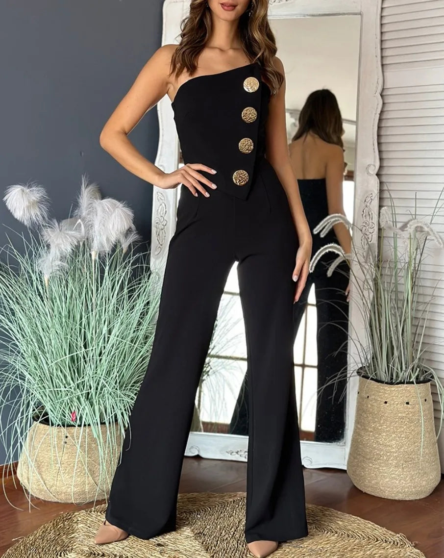 Woman wearing a black one-shoulder jumpsuit with gold buttons in a room with plants and a mirror.