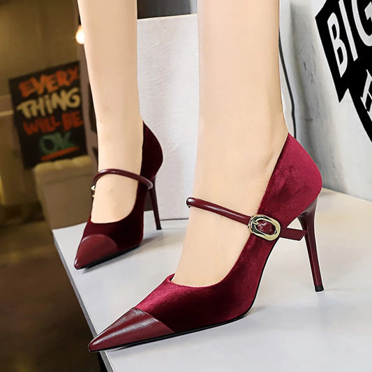 A pair of women's pointed-toe high heels in deep burgundy velvet with a smooth leather toe cap and a thin ankle strap with a gold-tone oval buckle, shown on a model's feet.
