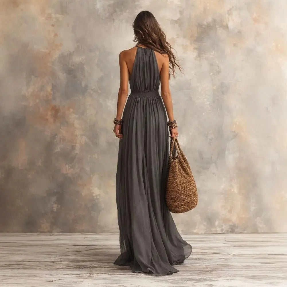 A woman stands with her back to the camera wearing a sleeveless, halter-neck, floor-length dark gray dress. She is holding a large woven straw tote bag against a textured, mottled studio backdrop with gray and beige tones.