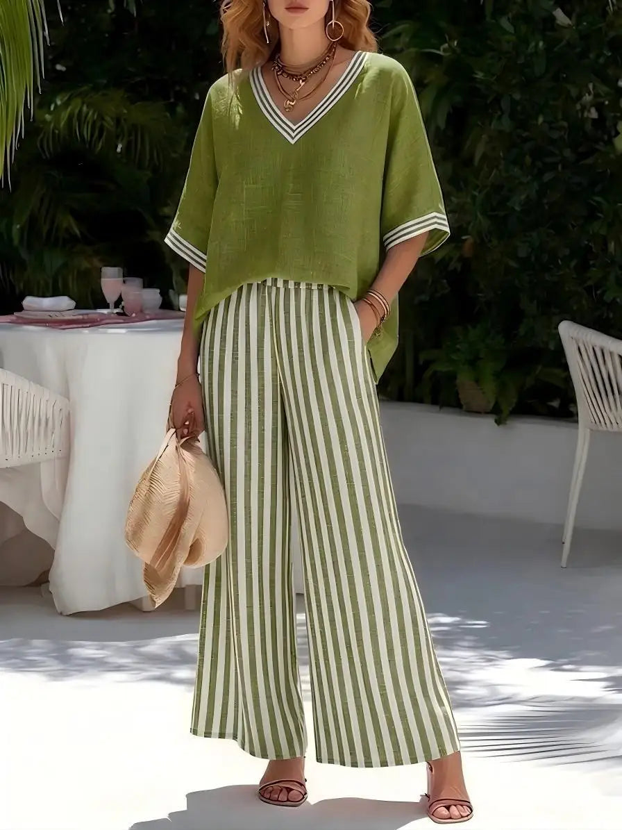 Woman in green top and striped pants standing outdoors with a table and chairs in the background.