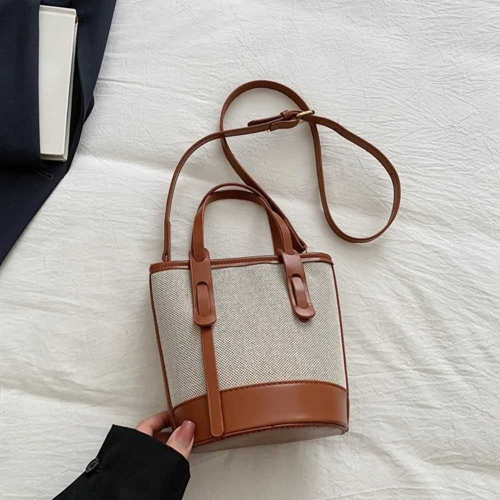 A small bucket-style handbag with a beige woven body and brown leather trim, shown on a white fabric surface. A hand in a black sleeve holds the lower left corner of the bag, and part of a dark blazer is visible at the upper left.