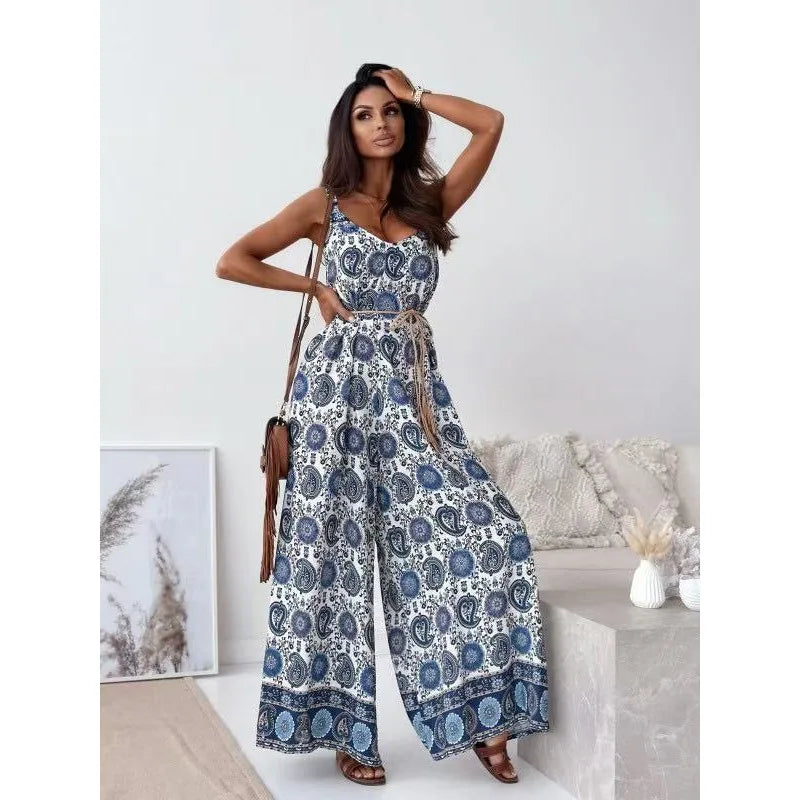 A woman models a sleeveless wide-leg jumpsuit with a blue and white paisley pattern and a matching tie belt, styled in a bright, modern room setting.