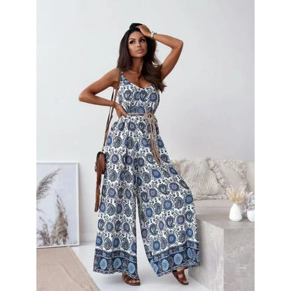 A woman models a sleeveless wide-leg jumpsuit with a blue and white paisley pattern and a matching tie belt, styled in a bright, modern room setting.