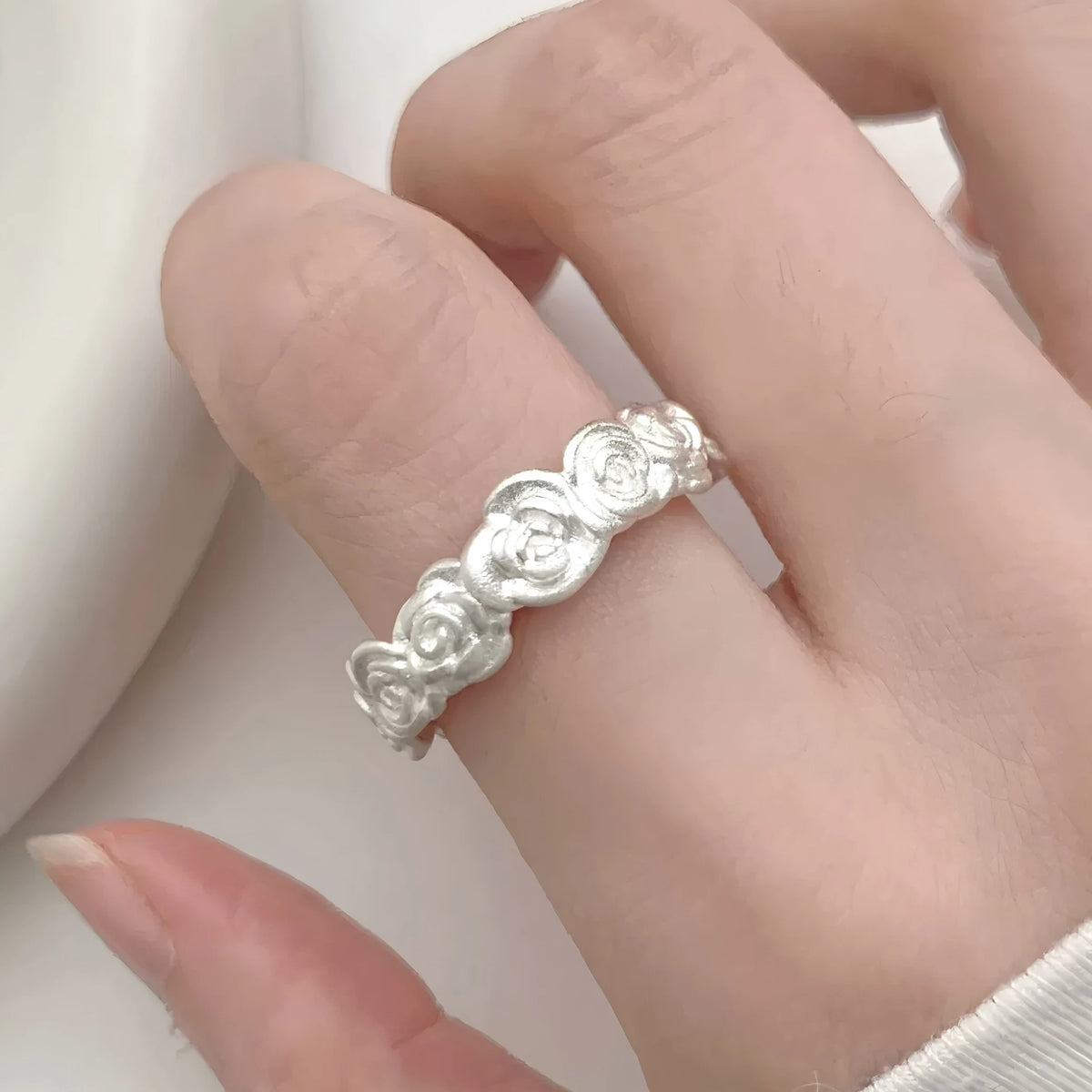 A close-up of a hand wearing a delicate silver ring with a continuous floral or rose-like textured design, set against a soft, light-colored background.