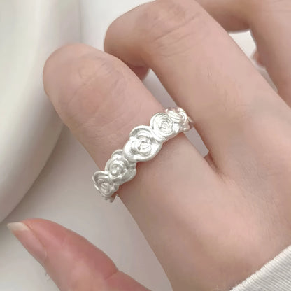 A close-up of a hand wearing a delicate silver ring with a continuous floral or rose-like textured design, set against a soft, light-colored background.