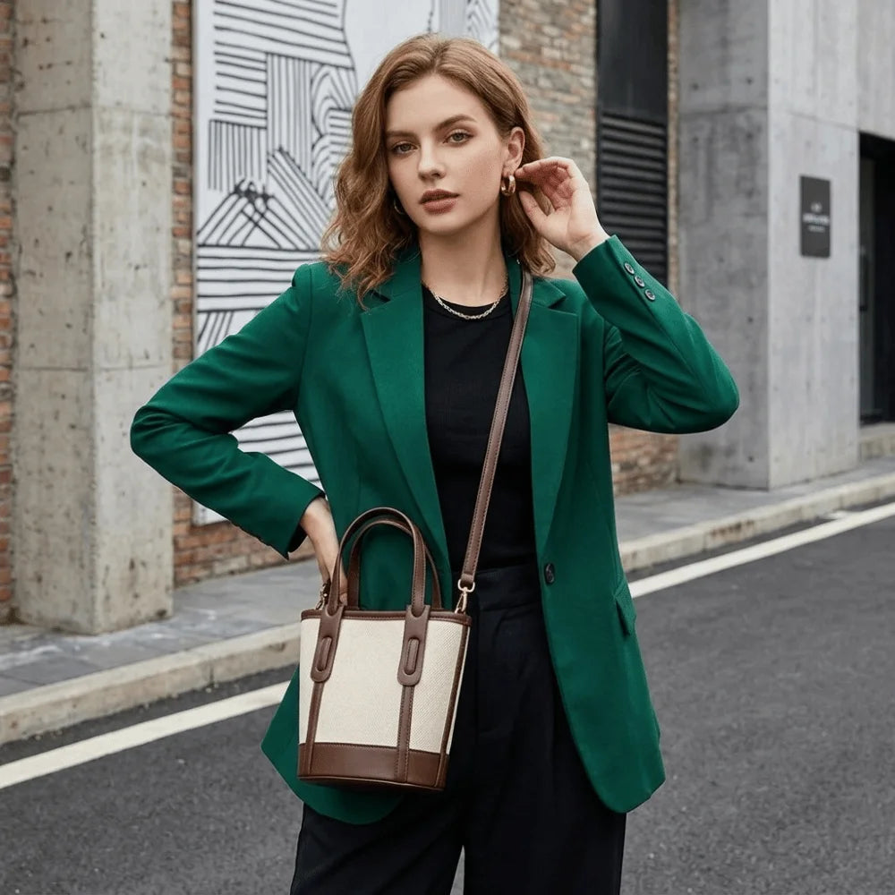 A woman stands on a city street wearing a bright green blazer, black outfit, and carrying a structured tote bag with cream panels and brown trim.