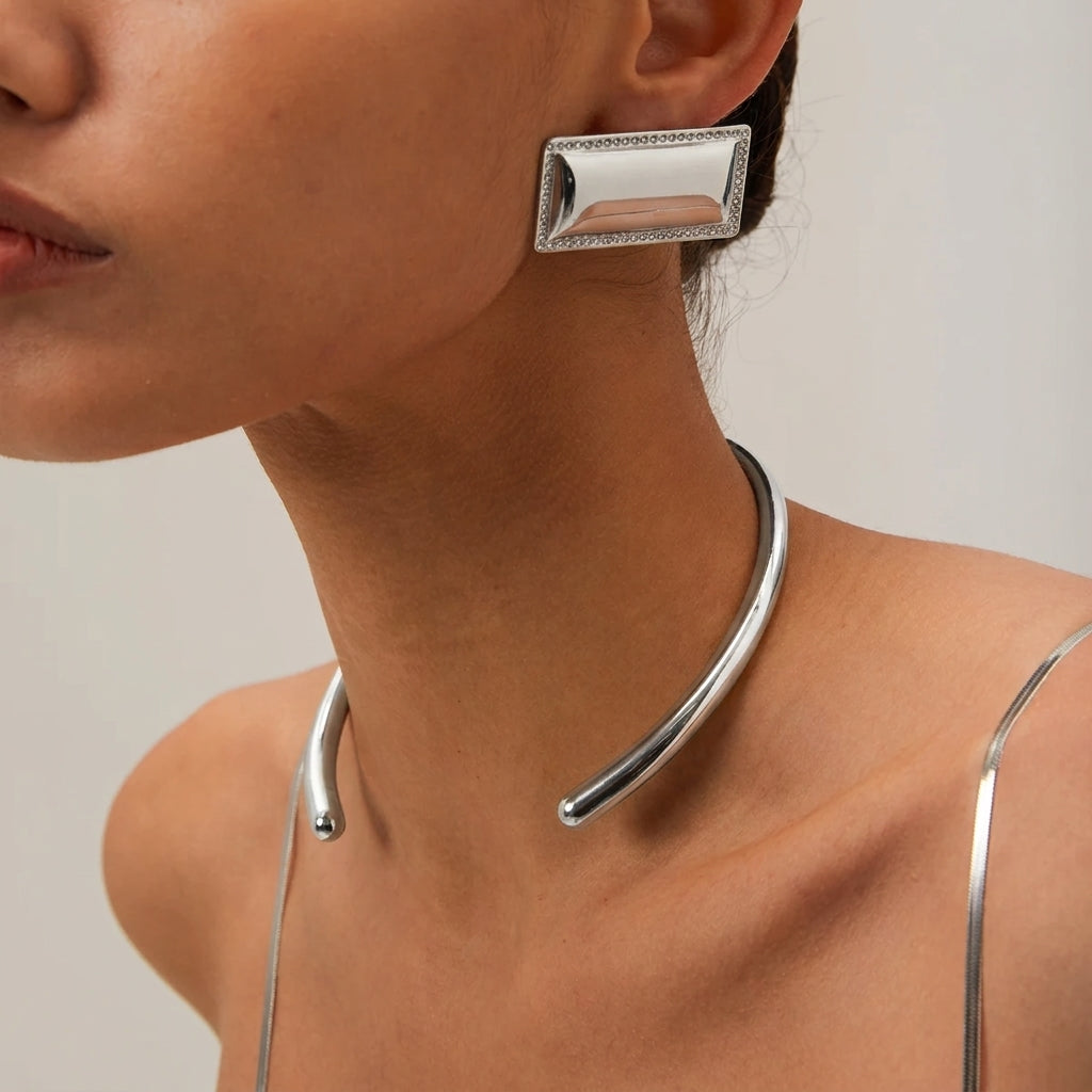 A close-up of a model wearing a polished silver-toned metal choker necklace and a matching rectangular stud earring with a beaded border.