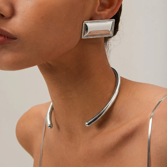 A close-up of a model wearing a polished silver-toned metal choker necklace and a matching rectangular stud earring with a beaded border.
