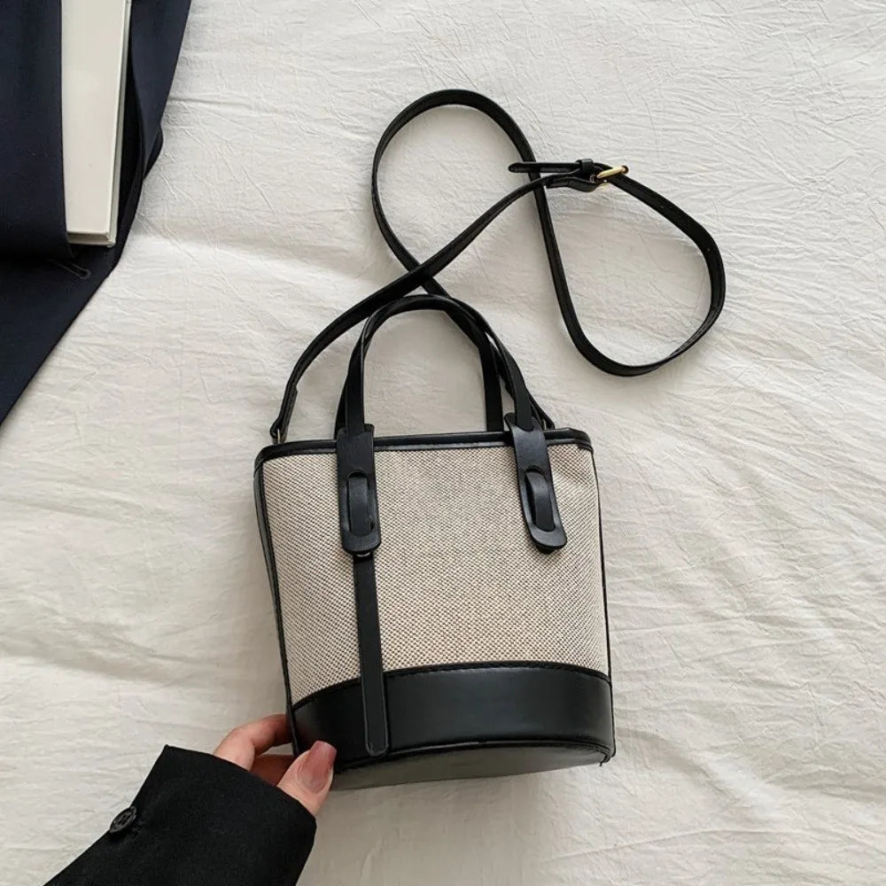 A bucket-style handbag with a beige woven body and black leather trim, handles, and shoulder strap, shown on a white fabric surface with a hand holding the sleeve of a black coat at the bottom left.