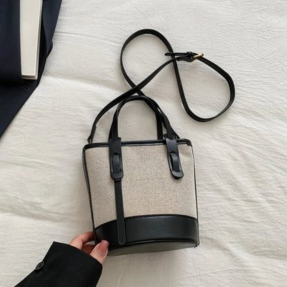 A bucket-style handbag with a beige woven body and black leather trim, handles, and shoulder strap, shown on a white fabric surface with a hand holding the sleeve of a black coat at the bottom left.