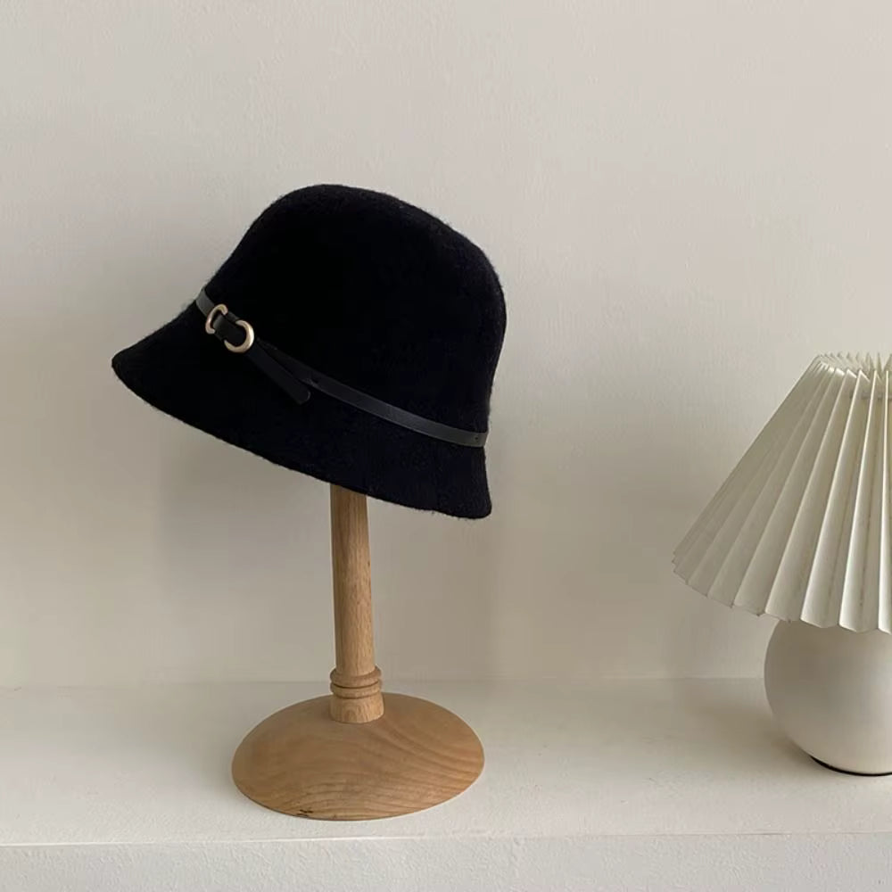 A black felt cloche hat with a black ribbon band and a small gold-tone buckle, displayed on a wooden stand against a light wall.