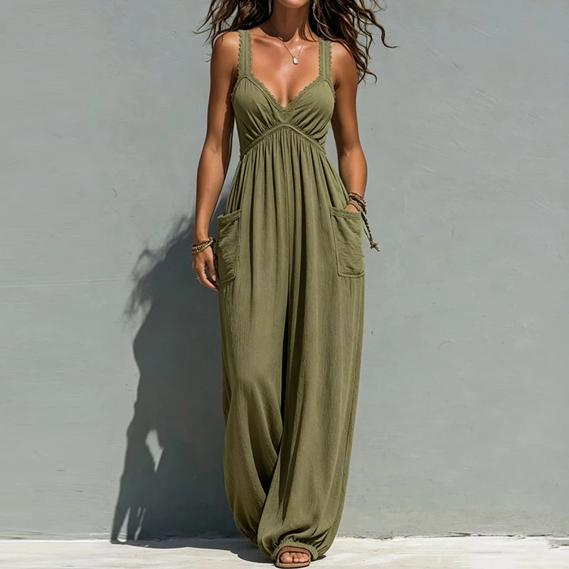 A woman models an olive green sleeveless maxi jumpsuit with a deep V-neckline, side pockets, and a loose, flowing silhouette, standing against a textured gray wall.
