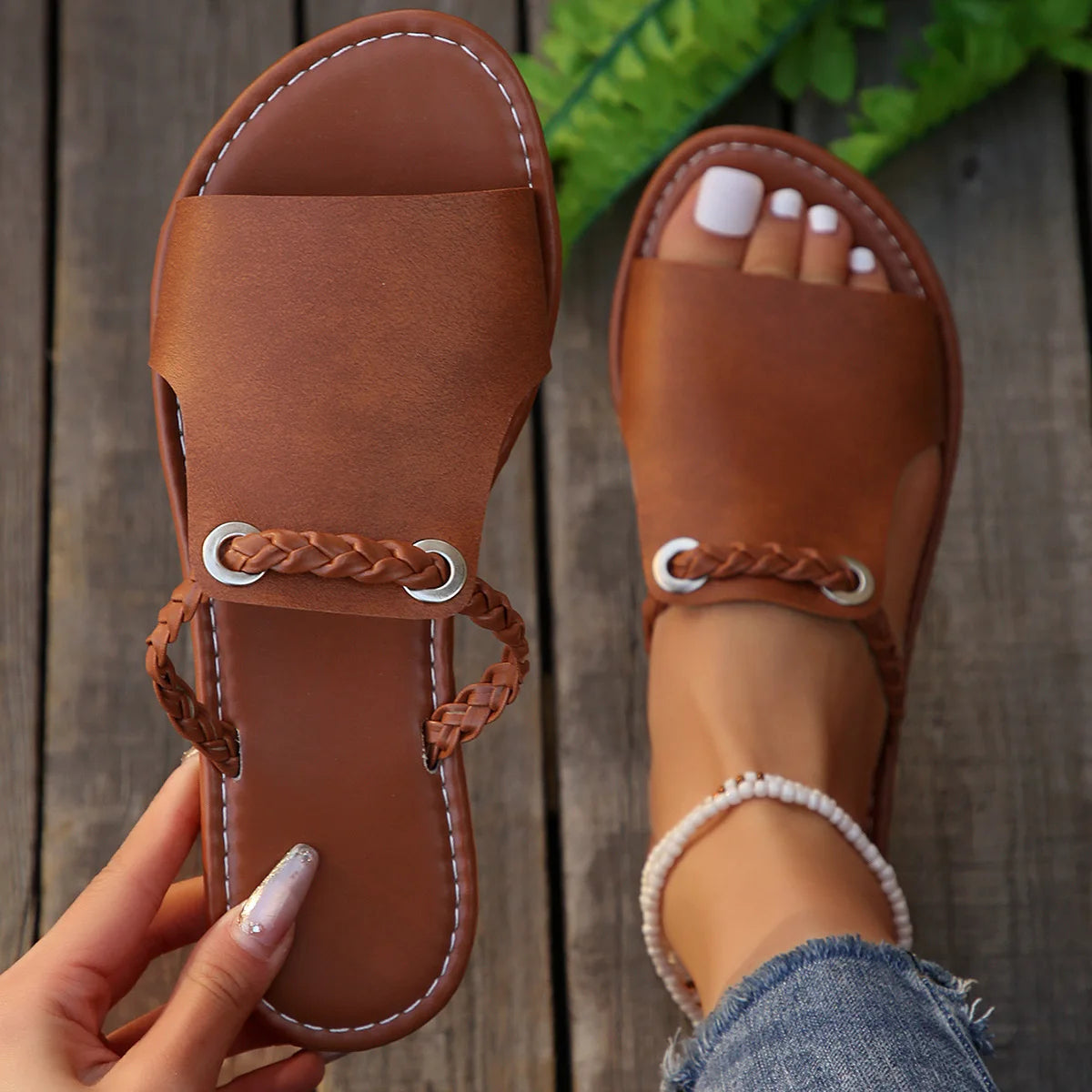 Brown leather sandals with braided straps held by a hand on a wooden floor.