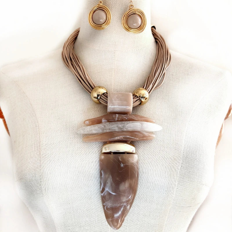 A statement necklace and matching drop earrings displayed on a cream-colored mannequin bust. The necklace features a multi-strand brown cord base, gold-toned metal beads, a rectangular taupe stone, and a large layered pendant with a teardrop-shaped brown agate-like centerpiece.
