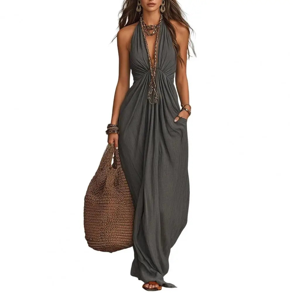A woman modeling a dark gray halter maxi dress with a deep plunging V-neckline, gathered waist, and side pockets, holding a large brown woven tote bag against a plain white background.