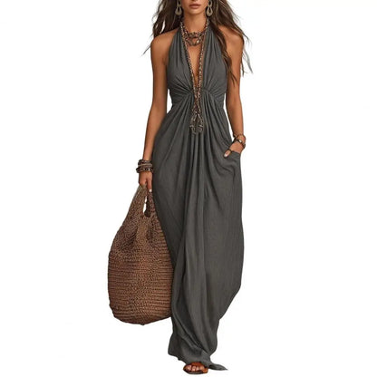 A woman modeling a dark gray halter maxi dress with a deep plunging V-neckline, gathered waist, and side pockets, holding a large brown woven tote bag against a plain white background.