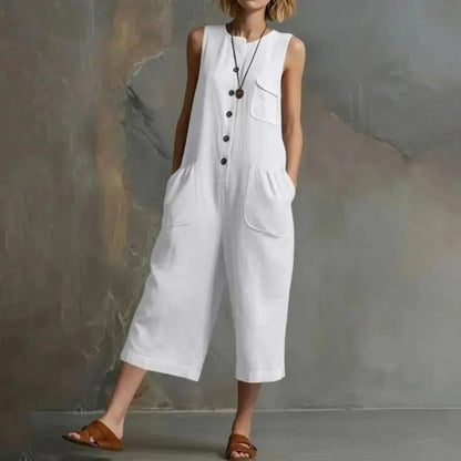 A woman wearing a white sleeveless linen-look jumpsuit with front buttons, chest pocket, and large patch pockets, standing against a textured gray wall. The brand name XJGOOD is printed in black in the upper left corner.