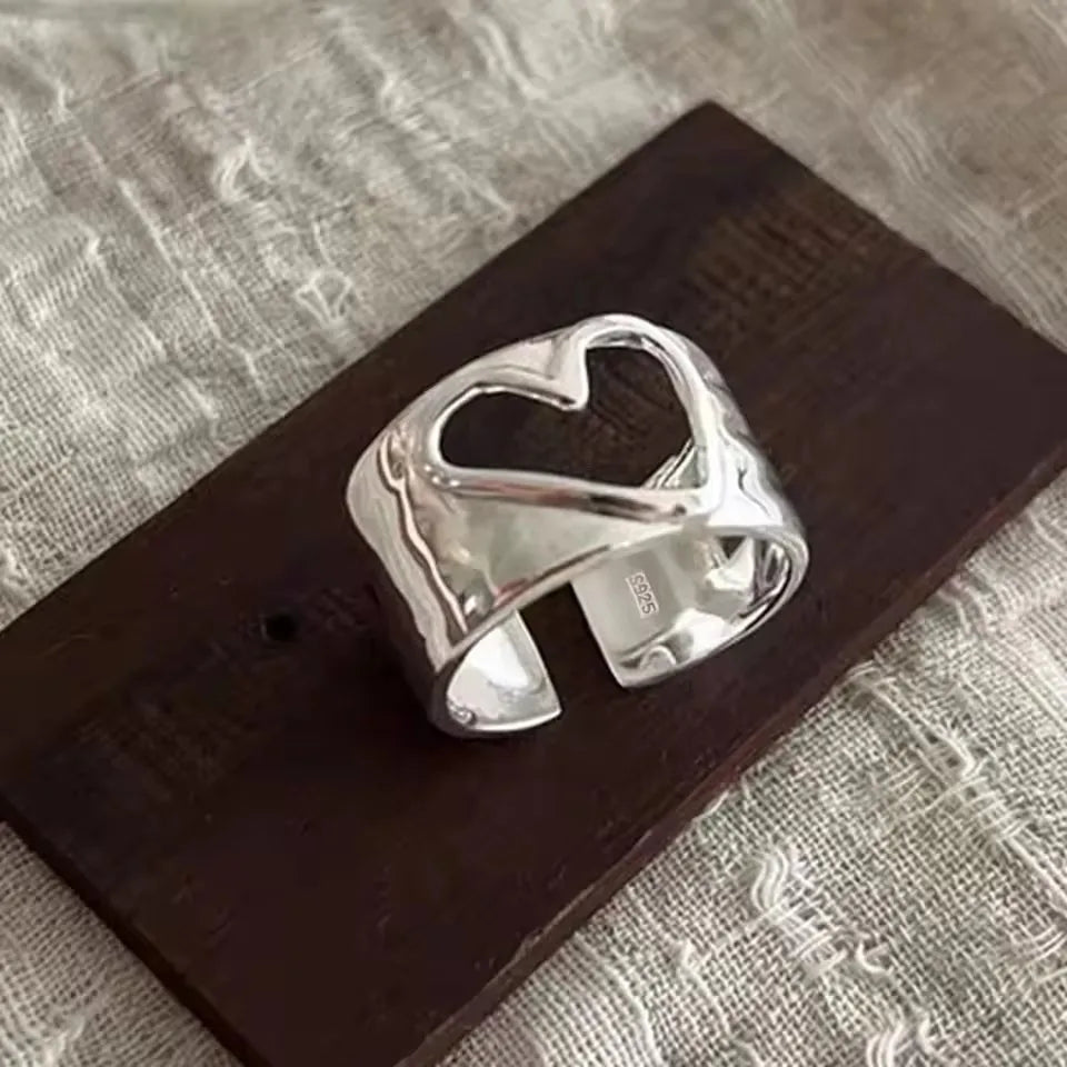 A polished silver cuff ring with a cutout heart design, displayed on a dark wooden block over a textured beige fabric background.