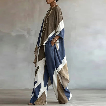 A woman models a loose, draped two-piece outfit with wide sleeves and wide-leg pants in a color-blocked abstract pattern of navy blue, taupe brown, cream, and white. She is standing in a studio with a textured gray background.
